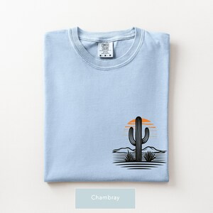 Puede incluir: Una camiseta azul claro con un gr&aacute;fico en la parte inferior derecha que muestra la silueta de un cactus negro, un sol naranja estilizado y l&iacute;neas horizontales que representan un paisaje des&eacute;rtico. La etiqueta interior dice Comfort Colors talla M.