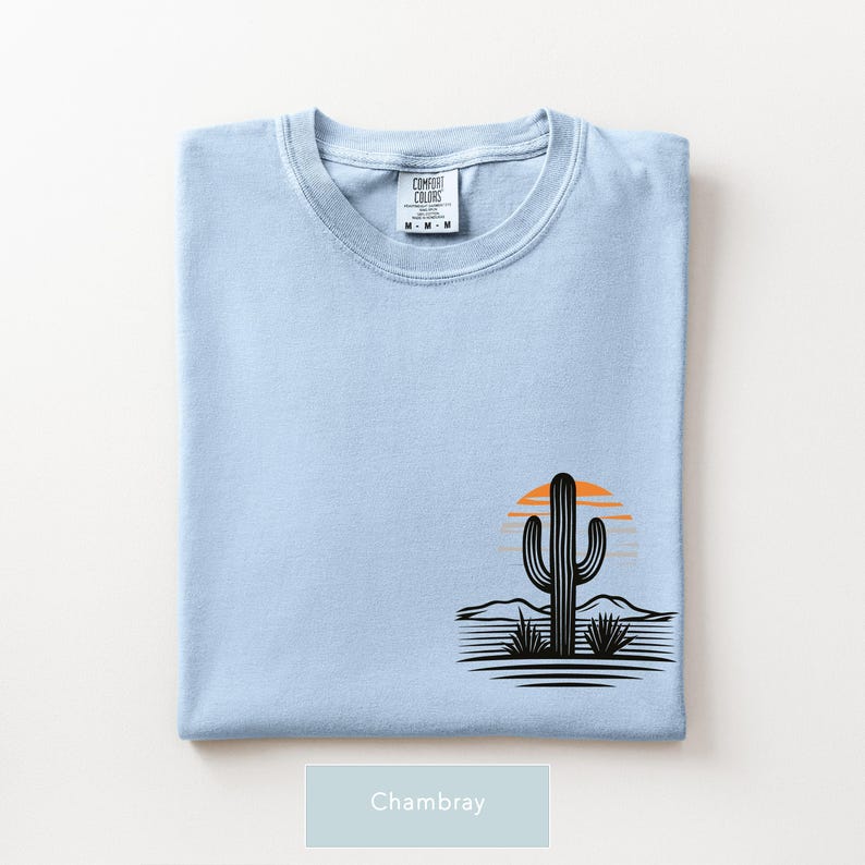 Puede incluir: Una camiseta azul claro con un gr&aacute;fico en la parte inferior derecha que muestra la silueta de un cactus negro, un sol naranja estilizado y l&iacute;neas horizontales que representan un paisaje des&eacute;rtico. La etiqueta interior dice Comfort Colors talla M.