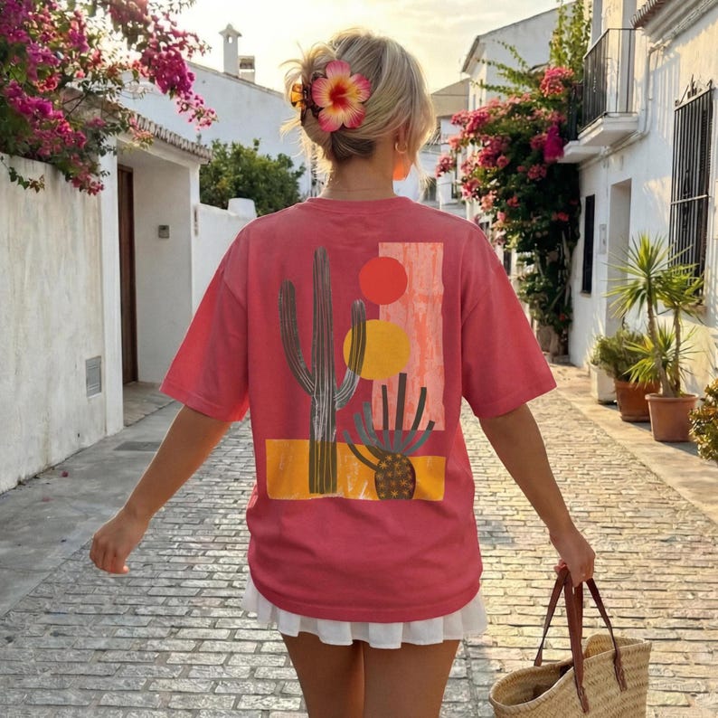 Puede incluir: Una camiseta de manga corta color coral con un estampado gr&aacute;fico en la espalda. El dise&ntilde;o incluye un cactus saguaro alto, un cactus redondo m&aacute;s peque&ntilde;o, un c&iacute;rculo rojo, un c&iacute;rculo amarillo y un bloque rectangular con un patr&oacute;n naranja texturizado.