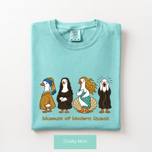 May include: A chalky mint t-shirt featuring a graphic of four ducks dressed as famous art figures. Below the ducks, the text reads "Museum of Modern Quack" in orange letters. The shirt has a Comfort Colors brand label at the inner neckline.