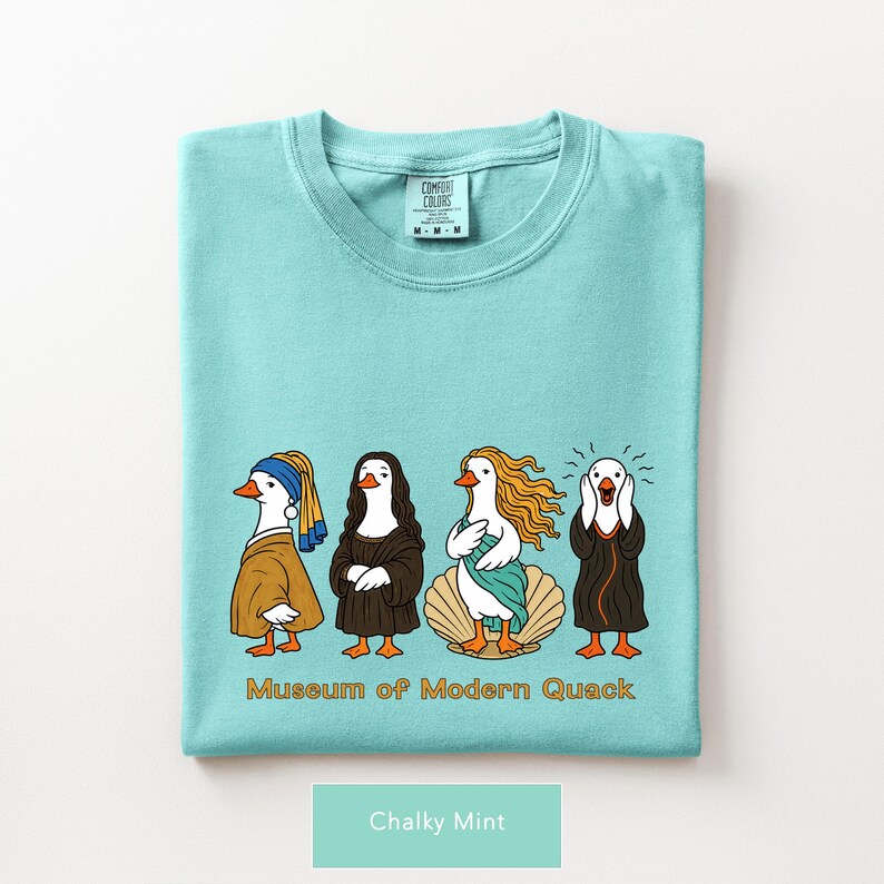May include: A chalky mint t-shirt featuring a graphic of four ducks dressed as famous art figures. Below the ducks, the text reads "Museum of Modern Quack" in orange letters. The shirt has a Comfort Colors brand label at the inner neckline.