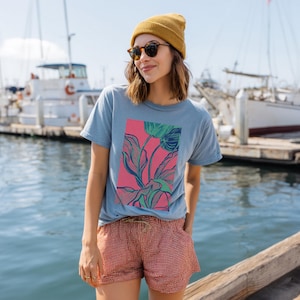 May include: A light blue t-shirt with a pink rectangle featuring a floral design in green and blue. The person is wearing a yellow beanie, sunglasses, and red and white checkered shorts. Boats are in the background.