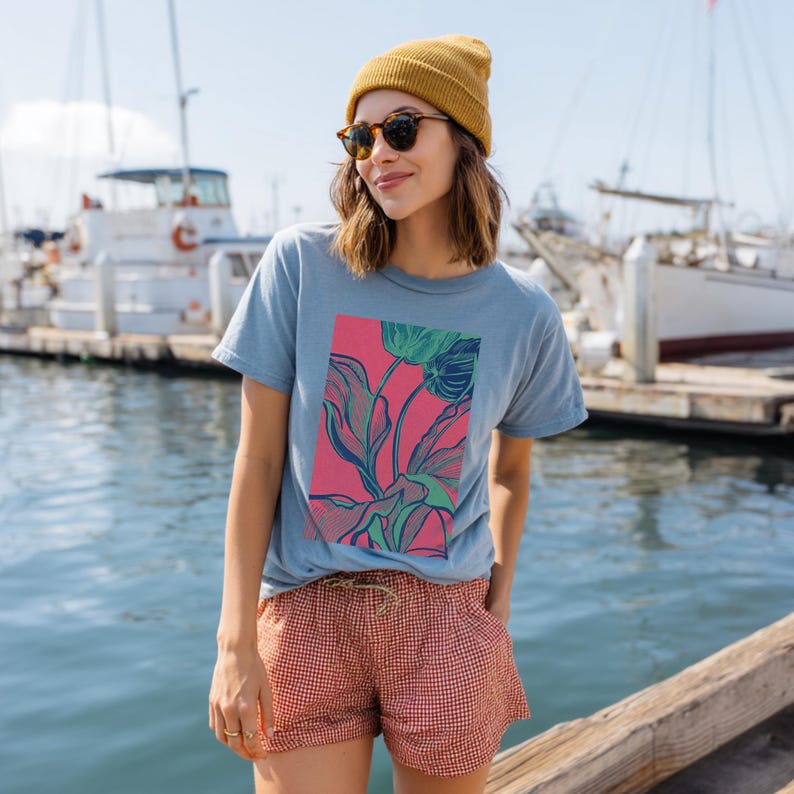 May include: A light blue t-shirt with a pink rectangle featuring a floral design in green and blue. The person is wearing a yellow beanie, sunglasses, and red and white checkered shorts. Boats are in the background.
