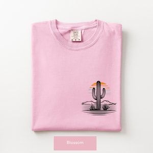 Puede incluir: Una camiseta rosa claro con un gr&aacute;fico en la parte inferior derecha. El dise&ntilde;o muestra la silueta negra de un cactus saguaro frente a un sol estilizado naranja y azul, con l&iacute;neas horizontales que representan un paisaje des&eacute;rtico. La etiqueta del cuello dice Comfort Colors.