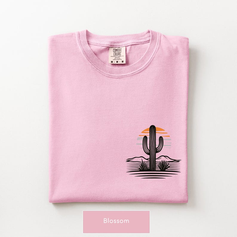 Puede incluir: Una camiseta rosa claro con un gr&aacute;fico en la parte inferior derecha. El dise&ntilde;o muestra la silueta negra de un cactus saguaro frente a un sol estilizado naranja y azul, con l&iacute;neas horizontales que representan un paisaje des&eacute;rtico. La etiqueta del cuello dice Comfort Colors.