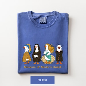 May include: A periwinkle blue t-shirt featuring a graphic of four ducks dressed as famous art figures. Below the ducks, the text reads "Museum of Modern Quack". The shirt has a crew neckline and a Comfort Colors brand label at the inner collar.