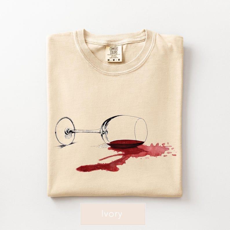 May include: Ivory-colored t-shirt featuring a graphic of a tipped-over wine glass with spilled red wine. The Comfort Colors brand label is visible at the top. The shirt is folded neatly.