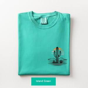 Puede incluir: Una camiseta color verde isla doblada con un peque&ntilde;o gr&aacute;fico en la parte inferior derecha. El gr&aacute;fico muestra la silueta negra de un cactus sobre un atardecer estilizado en naranja y azul claro, con l&iacute;neas horizontales del paisaje.
