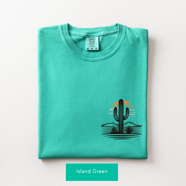 Puede incluir: Una camiseta color verde isla doblada con un peque&ntilde;o gr&aacute;fico en la parte inferior derecha. El gr&aacute;fico muestra la silueta negra de un cactus sobre un atardecer estilizado en naranja y azul claro, con l&iacute;neas horizontales del paisaje.