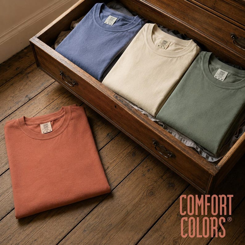 May include: Four crew neck t-shirts in solid colors including blue, cream, sage green, and terracotta. Each shirt features a Comfort Colors brand label at the inner neckline. The shirts are folded neatly, with three inside a wooden drawer and one placed outside.