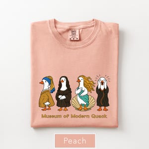 May include: A peach-colored t-shirt featuring a graphic of four ducks dressed as famous art figures. Below the ducks, the text reads "Museum of Modern Quack". The shirt has a crew neckline and a Comfort Colors brand label visible at the inner collar.