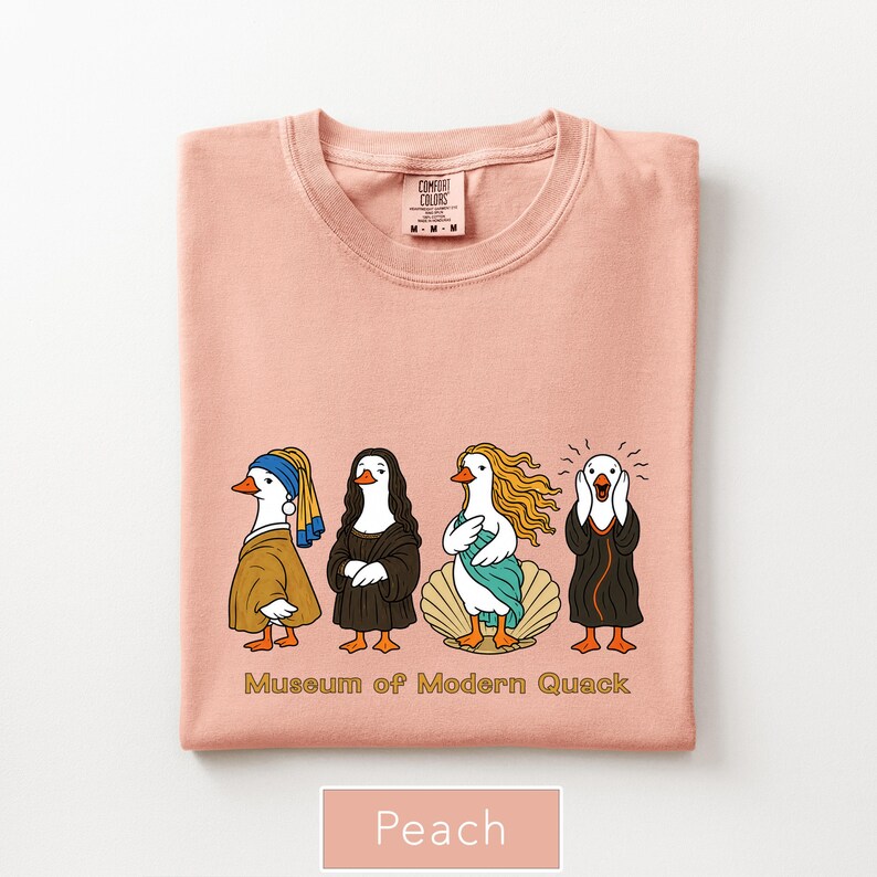 May include: A peach-colored t-shirt featuring a graphic of four ducks dressed as famous art figures. Below the ducks, the text reads "Museum of Modern Quack". The shirt has a crew neckline and a Comfort Colors brand label visible at the inner collar.