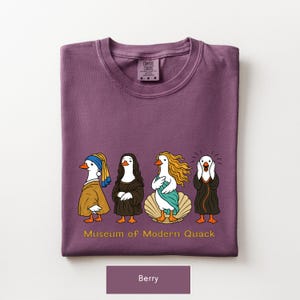 May include: A berry-colored t-shirt featuring a graphic of four ducks dressed as famous art figures. Below the ducks, the text reads "Museum of Modern Quack" in yellow. The shirt has a crew neck and a Comfort Colors brand label at the inner neckline.