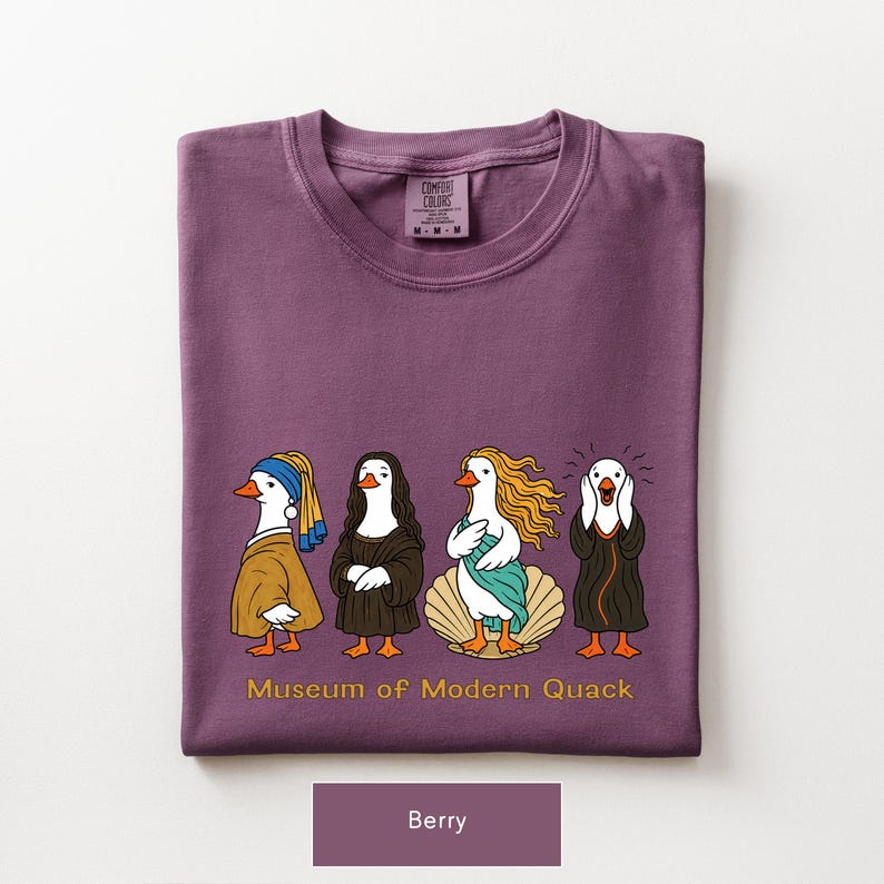 May include: A berry-colored t-shirt featuring a graphic of four ducks dressed as famous art figures. Below the ducks, the text reads "Museum of Modern Quack" in yellow. The shirt has a crew neck and a Comfort Colors brand label at the inner neckline.