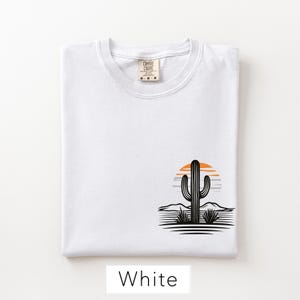 Puede incluir: Una camiseta blanca con un gr&aacute;fico en la parte inferior derecha. El gr&aacute;fico muestra la silueta de un cactus negro frente a un sol naranja y gris, con colinas des&eacute;rticas y l&iacute;neas de suelo negras. La etiqueta interior dice Comfort Colors M-M-M.