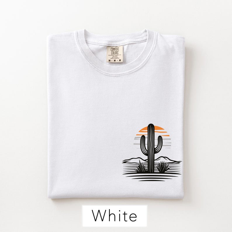 Puede incluir: Una camiseta blanca con un gr&aacute;fico en la parte inferior derecha. El gr&aacute;fico muestra la silueta de un cactus negro frente a un sol naranja y gris, con colinas des&eacute;rticas y l&iacute;neas de suelo negras. La etiqueta interior dice Comfort Colors M-M-M.