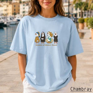 May include: A light blue t-shirt featuring a graphic print of four ducks dressed as famous art figures, including Girl with a Pearl Earring, Mona Lisa, The Birth of Venus, and The Scream, with the text Museum of Modern Quack below.