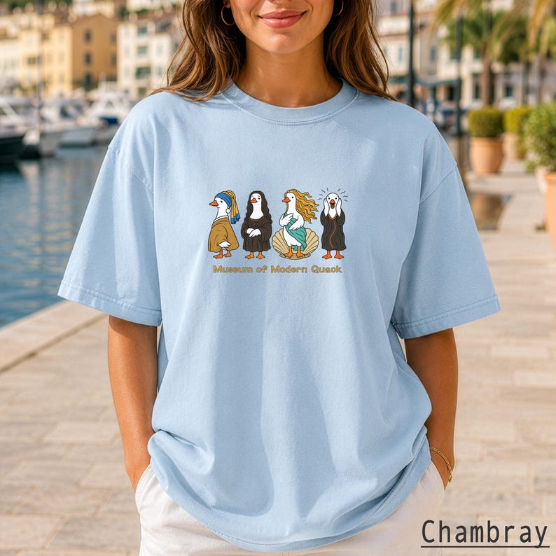 May include: A light blue t-shirt featuring a graphic print of four ducks dressed as famous art figures, including Girl with a Pearl Earring, Mona Lisa, The Birth of Venus, and The Scream, with the text Museum of Modern Quack below.