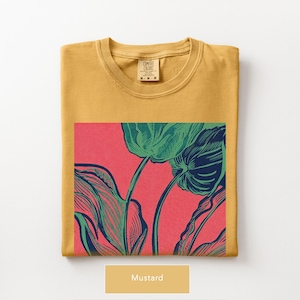 May include: Mustard-colored t-shirt with a large graphic of green leaves and stems on a red background. The shirt is folded neatly, with the Comfort Colors label visible. The word "Mustard" is displayed below the shirt.