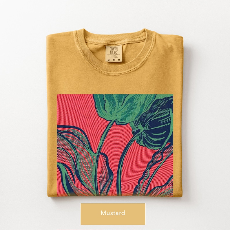 May include: Mustard-colored t-shirt with a large graphic of green leaves and stems on a red background. The shirt is folded neatly, with the Comfort Colors label visible. The word "Mustard" is displayed below the shirt.