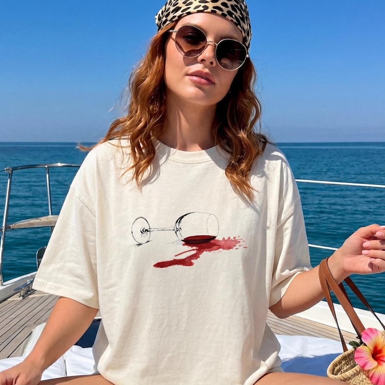 May include: An off-white short-sleeve t-shirt featuring a graphic illustration of a tipped-over wine glass with a red wine spill. The design is centered on the chest, showcasing a clear glass stem and a dark red liquid stain on the fabric.