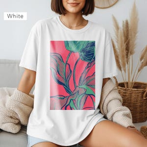 May include: White t-shirt featuring a vibrant floral design. The graphic showcases green and blue botanical illustrations against a bright pink background. The shirt is a casual, comfortable style.