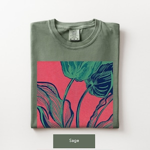 May include: Sage green t-shirt with a floral graphic. The graphic features a detailed illustration of green and blue flowers against a red background. The shirt is folded and has a tag that reads "Comfort Colors".