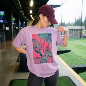 May include: A lilac-colored t-shirt featuring a vibrant floral design in shades of teal and green against a red background. The short-sleeved shirt is being held up by a person, showcasing the artwork.