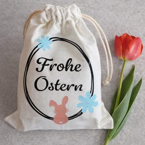 Könnte beinhalten: Ein cremefarbener Beutel mit Kordelzug und dem Text "Frohe Ostern" in einem schwarzen Kreis, verziert mit einem rosa Hasen und blauen Blumen. Eine rote Tulpe mit grünen Blättern liegt neben dem Beutel.