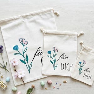 Könnte beinhalten: Drei cremefarbene Kordelzugbeutel mit Blumendesigns und dem Text "für Dich". Die Blumen sind blau und rosa mit grünen Stielen. Die Beutel sind in verschiedenen Größen gehalten und haben eine natürliche, rustikale Ästhetik.