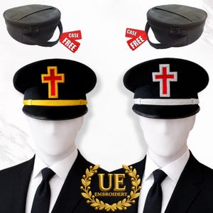 May include: Two black hats with a cross emblem, one with a gold band and the other with a white band. The hats are on mannequins wearing black suits and ties. Above the hats are two black cases. The text "UE EMBROIDERY" is in a gold wreath.