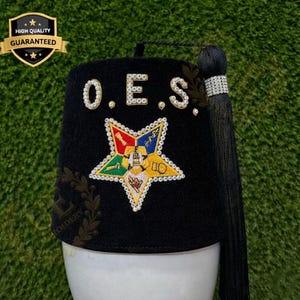 Puede incluir: Gorra fez negra con las letras "O.E.S." en blanco, adornada con pedrería. Un emblema de estrella colorida está centrado en la parte delantera. Una borla negra cuelga de un lado. El sombrero se muestra sobre una forma blanca sobre un fondo verde.