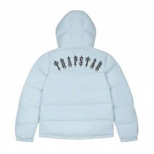 以下が含まれることがあります： フード付きのライトブルーのパファージャケット。「TRAPSTAR」の文字が、背面上部に様式化されたフォントで刺繍されています。ジャケットはキルティングデザインで、フルレングスのジッパーが付いています。