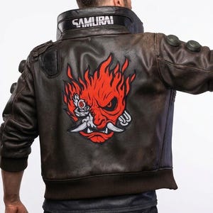 Pode incluir: Casaco de couro castanho com um gráfico vermelho e branco nas costas. O casaco tem a palavra "SAMURAI" na gola. O casaco tem um estilo bomber com bainha e punhos canelados. O casaco é usado por uma pessoa.