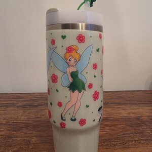 Puede incluir: Un vaso brillante de color claro con una imagen de dibujos animados de Campanilla con flores rosas y corazones verdes. El vaso tiene una tapa blanca, una pajita verde y una banda plateada. El dise&ntilde;o se repite.