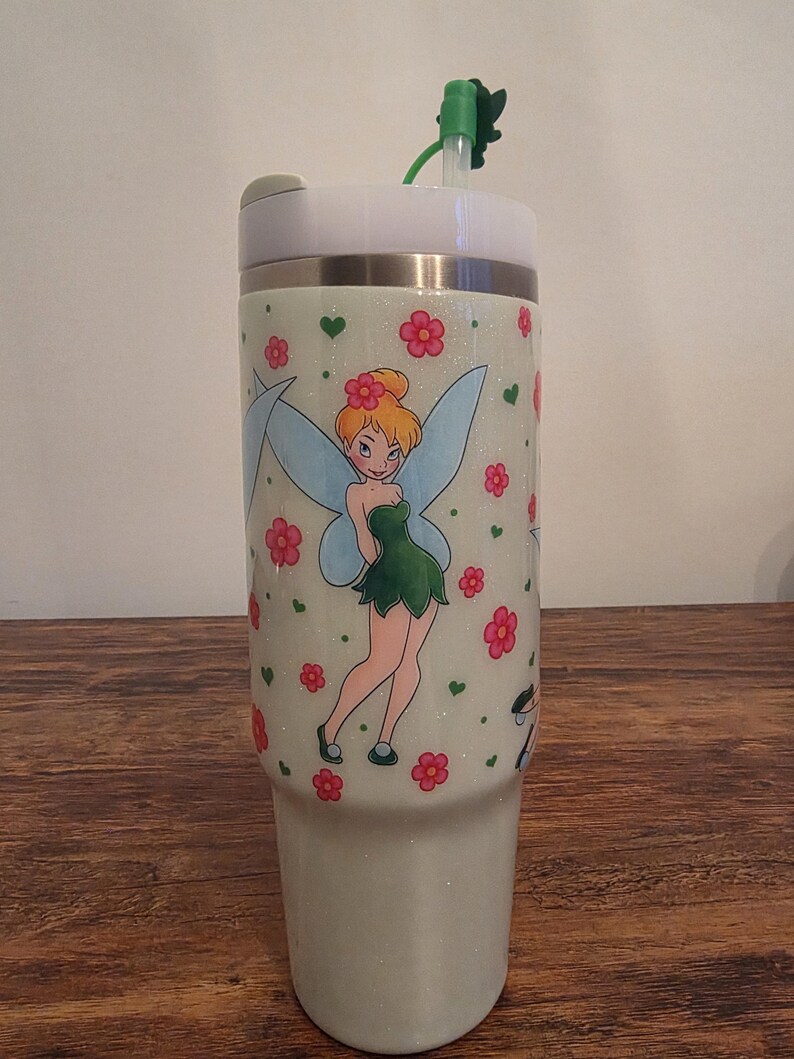 Puede incluir: Un vaso brillante de color claro con una imagen de dibujos animados de Campanilla con flores rosas y corazones verdes. El vaso tiene una tapa blanca, una pajita verde y una banda plateada. El dise&ntilde;o se repite.