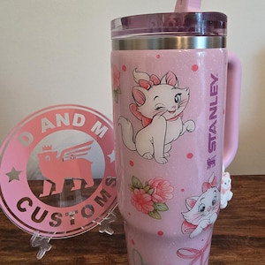 May include: A light pink glittery Stanley tumbler featuring Marie the cat from The Aristocats, pink polka dots, floral accents, and pink bows. The tumbler has a clear lid with a pink straw and a handle. Beside it is a round pink logo sign reading D AND M CUSTOMS.