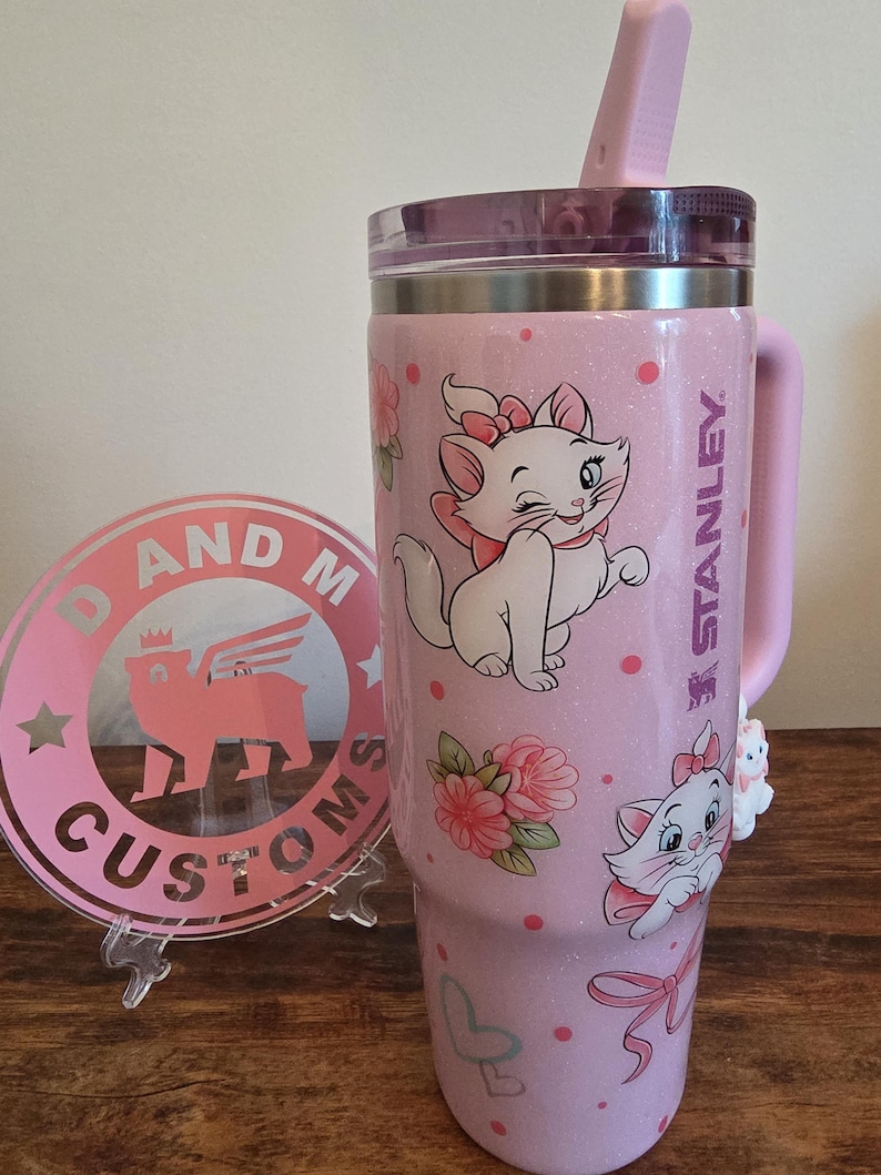 May include: A light pink glittery Stanley tumbler featuring Marie the cat from The Aristocats, pink polka dots, floral accents, and pink bows. The tumbler has a clear lid with a pink straw and a handle. Beside it is a round pink logo sign reading D AND M CUSTOMS.