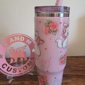 May include: A light pink glitter tumbler with a clear lid and a pink straw. The tumbler features a repeating pattern of white cartoon cats with pink bows, pink flowers with green leaves, small pink dots, and outlined hearts in pink and light blue.