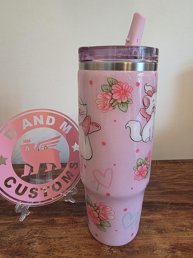May include: A light pink glitter tumbler with a clear lid and a pink straw. The tumbler features a repeating pattern of white cartoon cats with pink bows, pink flowers with green leaves, small pink dots, and outlined hearts in pink and light blue.