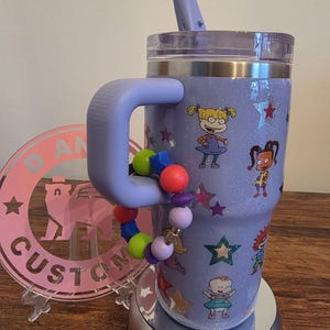 Puede incluir: Un vaso con purpurina lavanda con asa, con calcoman&iacute;as de personajes de dibujos animados y detalles de estrellas. Una pulsera de cuentas de colores est&aacute; unida al asa. El vaso tiene tapa y pajita a juego.