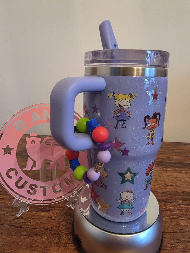 Puede incluir: Un vaso con purpurina lavanda con asa, con calcoman&iacute;as de personajes de dibujos animados y detalles de estrellas. Una pulsera de cuentas de colores est&aacute; unida al asa. El vaso tiene tapa y pajita a juego.