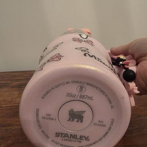May include: Pink Stanley tumbler with a Minnie Mouse theme. The tumbler has a glittery finish and features Minnie Mouse graphics. The bottom of the tumbler has the Stanley brand logo and text that reads "30oz / 887mL".