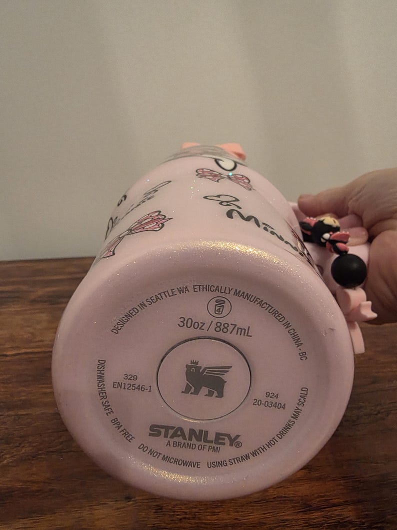 May include: Pink Stanley tumbler with a Minnie Mouse theme. The tumbler has a glittery finish and features Minnie Mouse graphics. The bottom of the tumbler has the Stanley brand logo and text that reads "30oz / 887mL".
