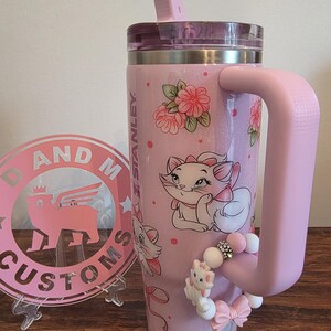 May include: A light pink Stanley tumbler with a glitter finish, featuring Marie the cat from The Aristocats, pink floral accents, and a pink straw. A beaded charm with a small Marie figure and a pink bow is attached to the large side handle.