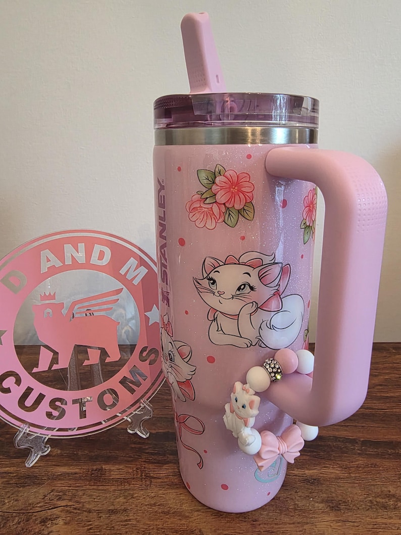 May include: A light pink Stanley tumbler with a glitter finish, featuring Marie the cat from The Aristocats, pink floral accents, and a pink straw. A beaded charm with a small Marie figure and a pink bow is attached to the large side handle.