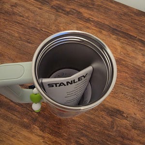 Puede incluir: Un termo de viaje Stanley de acero inoxidable con un asa verde claro. El interior del termo es gris oscuro y hay un manual de instrucciones plegado en su interior. Un peque&ntilde;o adorno verde y blanco cuelga del asa. El termo est&aacute; sobre una superficie de madera.