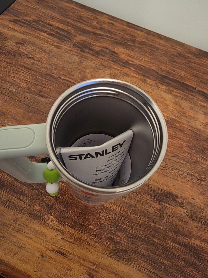 Puede incluir: Un termo de viaje Stanley de acero inoxidable con un asa verde claro. El interior del termo es gris oscuro y hay un manual de instrucciones plegado en su interior. Un peque&ntilde;o adorno verde y blanco cuelga del asa. El termo est&aacute; sobre una superficie de madera.