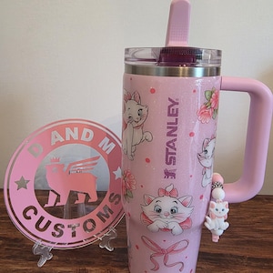 May include: A light pink glittery Stanley tumbler featuring Marie the cat from The Aristocats, floral accents, and a pink bow. A matching pink circular acrylic sign with the text D AND M CUSTOMS and a winged cat logo stands beside it.