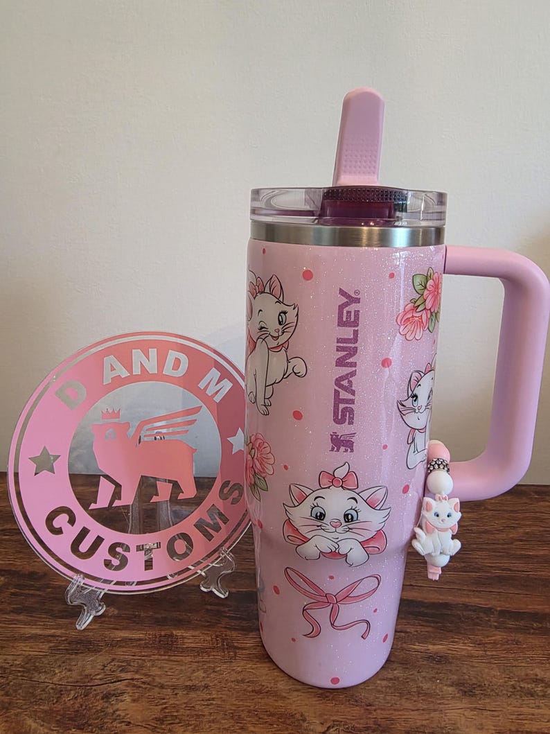 May include: A light pink glittery Stanley tumbler featuring Marie the cat from The Aristocats, floral accents, and a pink bow. A matching pink circular acrylic sign with the text D AND M CUSTOMS and a winged cat logo stands beside it.